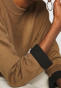 Pull marron avec un col côtelé et des poignets noirs. Le tissu semble doux, présentant une coupe ample avec des manches froncées au niveau des coudes.