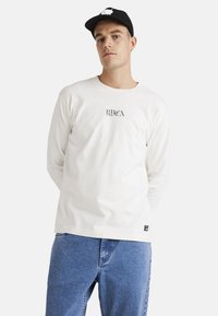 White long-sleeve shirt with black graphic text on the front, paired with blue denim jeans. The model wears a black cap.