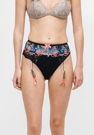 Žena u bež čipkastom grudnjaku i crnim gaćicama visokog struka s cvjetnim ružičasto-plavim vezom i ružičastim vrpcama, stoji na bijeloj pozadini.