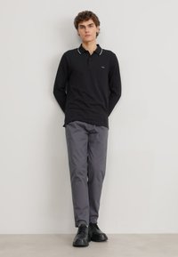 Polo a maniche lunghe nera con dettagli del colletto bianco, abbinato a pantaloni grigi e scarpe nere. Il modello è in piedi con le mani dietro la schiena su uno sfondo neutro.