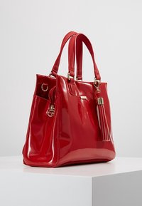 Borsa in pelle rossa lucida con una forma strutturata, doppio manico, un accento in metallo dorato e un attaccamento di nappa decorativa.