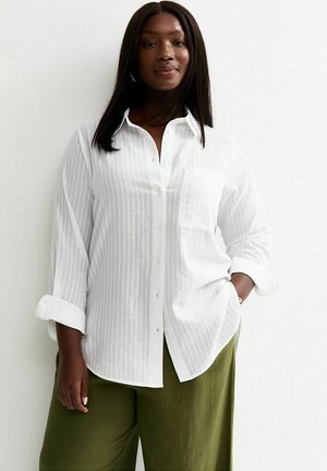 Vrouw draagt een wit overhemd met opgerolde mouwen en olijfgroene broek, staand tegen een effen witte achtergrond.