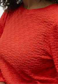 Rød tekstureret bluse med et subtilt vævet mønster, rund halsudskæring og let puffede ærmer. Stoffet ser let og åndbart ud.