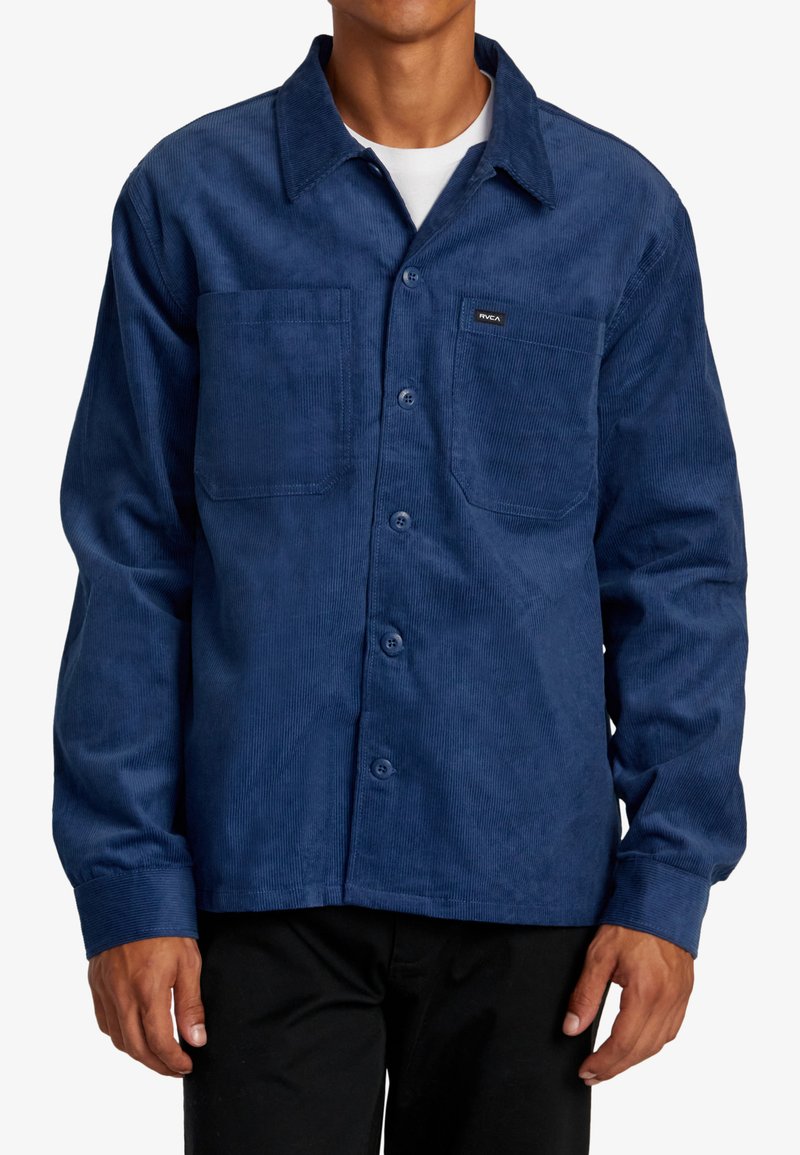 Camicia in corduroy blu con chiusura a bottoni, due tasche sul petto e un'etichetta. Presenta un colletto a punta e maniche lunghe.