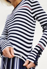 Pruhovaný svetřík z úpletu v námořnické modři a bílé barvě s výstřihem do V, zdobený červeným dekorativním šitím. Dlouhé rukávy s manžetami na knoflíky a žebrovanou texturou.