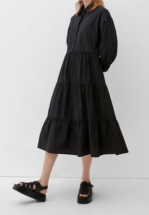 Woman wearing a long black tiered dress with balloon sleeves and black platform sandals standing against a plain background.