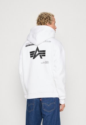 Persoon met krullend blond haar die een witte hoodie draagt met een zwart Alpha Industries-logo en tekst, gecombineerd met een blauwe spijkerbroek, gezien van achteren.