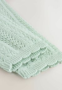 Adīta gaiši piparmētru zaļa auduma ar teksturētu rakstu, kuram ir viļņveida malas un ribota apdare apakšmalā.