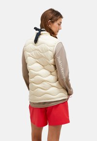 Gilet beige rembourré avec un design en quilting ondulé, associé à une chemise à manches longues marron texturée et un short rouge. Écharpe noire nouée au cou.