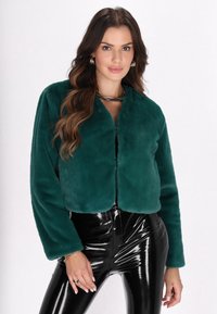 Teal faux fur jakke med cropped design og brede ærmer, parret med højt shine sorte leggings, der fremhæver en tætsiddende silhuet.