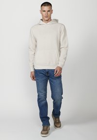Felpa beige con cappuccio, tasca frontale e logo ricamato; abbinata a jeans blu e scarpe beige con dettagli marroni.