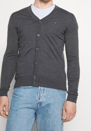 Uomo che indossa un cardigan grigio scuro abbottonato sopra una maglietta bianca e jeans azzurri, in piedi davanti a uno sfondo semplice.