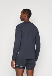 Camisola atlética de mangas longas em azul escuro, feita de tecido leve com costuras planas. Apresenta um decote redondo e um painel traseiro texturizado.