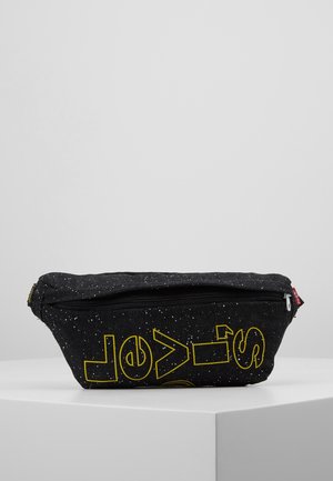 Sac banane en tissu noir tacheté avec texte contouré jaune et une fermeture éclair sur le dessus, présenté sur un socle blanc sur fond gris.
