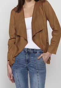 Femme portant une veste marron ouverte à revers drapés, par-dessus un haut blanc et un jean bleu taille haute, main dans la poche.