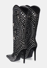 Botas negras hasta la rodilla con punta afilada, tacón alto de aguja y decoradas con un patrón de tachuelas plateadas y piedras de imitación.