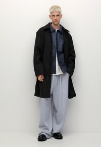 Schwarzer langer Mantel über einem blauen Jeanshemd, einem weißen T-Shirt und grauen Jogginghosen. Schwarzes Schuhwerk. Das Modell steht vor einem weißen Hintergrund.