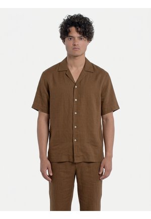 Giovane uomo con capelli ricci che indossa una camicia marrone a maniche corte con bottoni e pantaloni abbinati, in piedi davanti a uno sfondo bianco semplice.