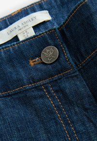 Tumši zili džinsi ar sudraba metāla pogu, kurai ir reljefs zieda dizains. Šuves ir kontrastējošā zelta krāsā.