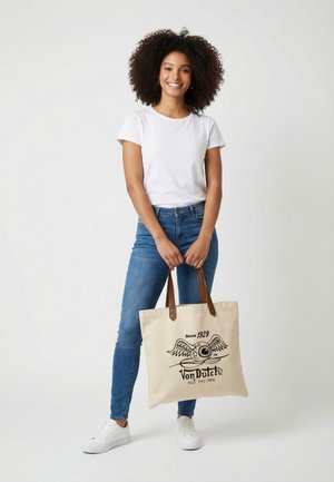 Donna con capelli ricci che indossa una t-shirt bianca e jeans blu, che tiene una borsa beige con logo nero di occhio alato e testo "Von Dutch."