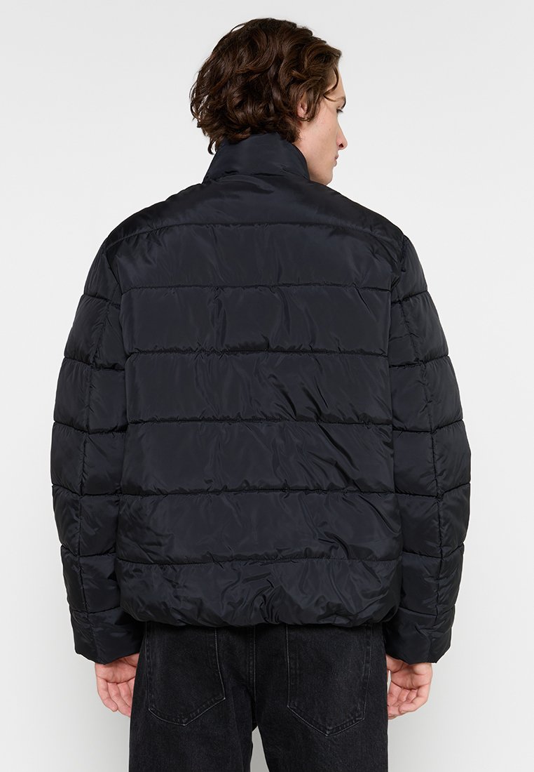 Chaqueta de plumas negra con cuello alto, costuras horizontales, puños elásticos y un corte ligeramente recortado, hecha de material sintético ligero.