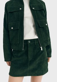 Chaqueta de pana verde con dos bolsillos frontales y herrajes plateados, combinada con una falda a juego. Ambos tienen una textura de canalé vertical.