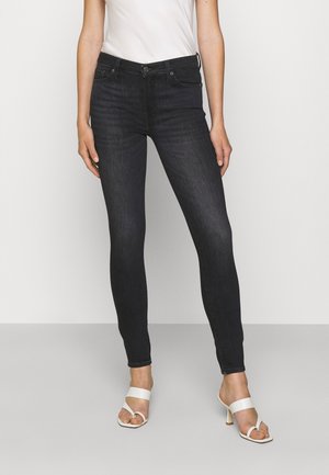 Femme portant un jean skinny noir, un top blanc sans manches et des sandales blanches à talons, debout devant un fond clair uni.