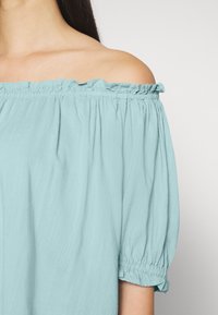 Blusa azul clara, off-the-shoulder, feita de tecido suave, com decote franzido e mangas balão elásticas, apresentando um corte descontraído.
