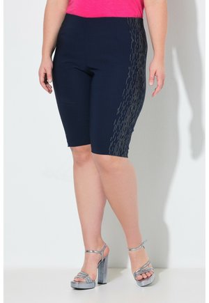 Frau trägt marineblaue kniehohe Shorts mit seitlichen Steppmuster, pinkes Oberteil und silberne Sandalen mit hohen Absätzen, steht vor einer weißen Wand.