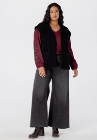 Zwarte textuurvest met twee voorzakken over een bordeauxrode langemouwen top, gedragen met grijze wijde broek en bruine enkellaarsjes met hak.