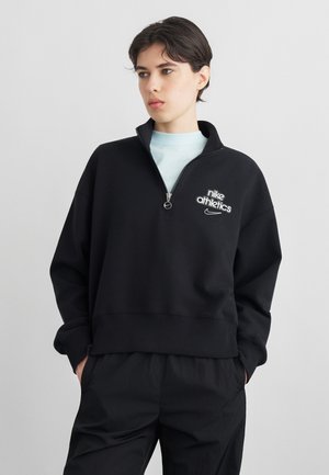 Osoba o krótkich ciemnych włosach, ubrana w czarną bluzę sportową Nike Athletics z zamkiem do połowy oraz czarne spodnie, z rękami w kieszeniach, patrząca na bok.