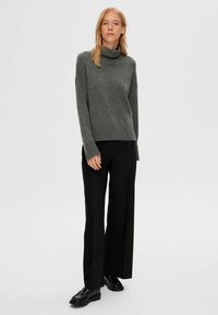 Grau gestrickter Pullover mit lässigem Schnitt, ausgestattet mit einem strukturierten Strickmuster und langen Ärmeln, kombiniert mit schwarzen weiten Hosen und schwarzen Schuhen.