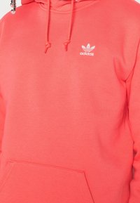Korallrosa Adidas-hoodie med frontficka, dragsko och en liten vit logotyp på bröstet, tillverkad av mjukt tyg.