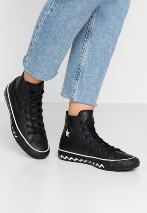 Osoba nosząca czarne, wysokie trampki z białym, zygzakowatym detalem podeszwy i emblematem gwiazdy, w połączeniu z jasnoniebieskimi, skróconymi dżinsami.