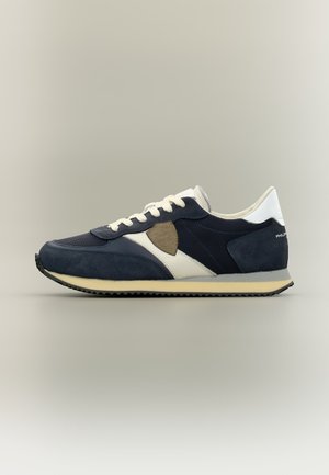 Baskets bleu marine et blanches avec des détails en daim, des lacets blancs, une semelle beige et une pièce sombre sur le côté, présentées sur un fond neutre.