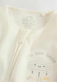 Otroški bodi kremaste barve z mehko teksturo, z motivom oblaka in vezenim napisom »sem nov tukaj«. Zapira se s sredinskim pritisnim zapiranjem.