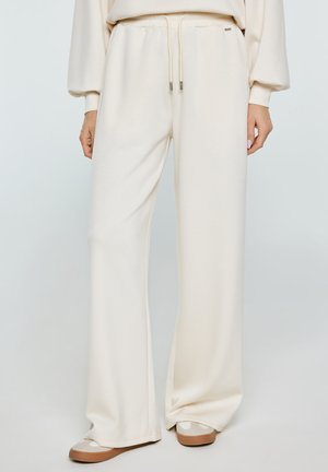 Kobieta w kremowych szerokich spodniach dresowych z wiązanym w pasie sznurkiem oraz beżowych sportowych butach stoi na jednolitym tle.