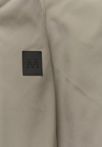 Beige tygsektion med en texturerad svart rektangulär fläck med ett präglat 'M'-logotyp. Slät, matt yta med subtil lyster.