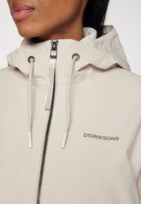 Veste imperméable beige avec capuche, fermeture éclair noire contrastante, cordons de serrage ajustables et logo "DIDRIKSONS" sur la poitrine. Texture lisse.