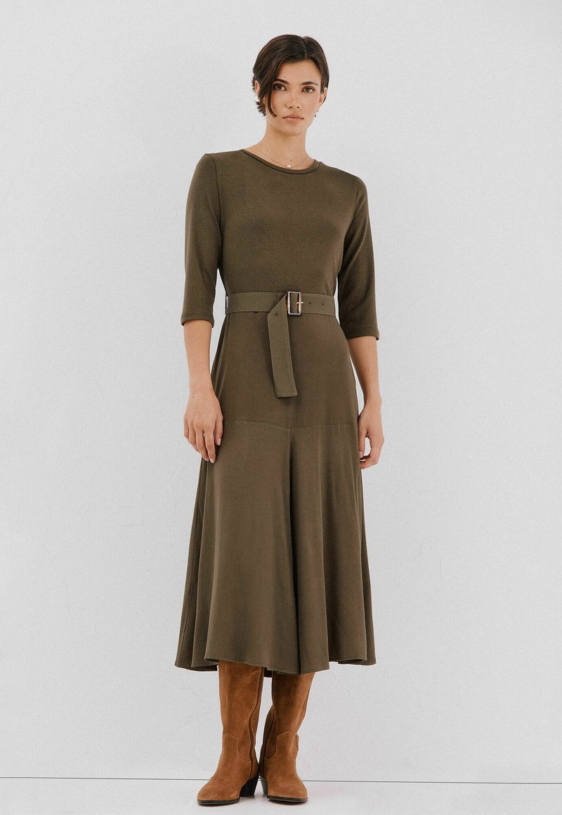 Oliwkowa sukienka do kolan z rękawami o długości trzech ćwierci, dopasowanym talią i pasującym paskiem; w zestawieniu z brązowymi, wysokimi do kolan butami.