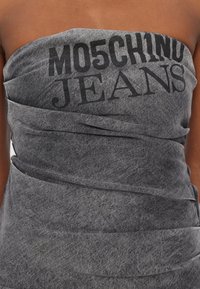 Sivi traper bez naramenica vrh s modernim omotanjem i istaknutim crnim natpisom "MOSCHINO JEANS" sprijeda. Tekstura koja se proteže cijelim proizvodom.