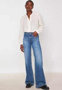 Donna che indossa una camicia bianca a bottone, jeans blu a vita alta e gamba larga, mocassini neri e orecchini a cerchio dorati, in piedi su uno sfondo neutro.