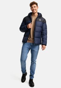 Marineblå pufferjakke med grå hætte og skulderpaneler, lynlås-lukning og to lommer. Båret over en tan sweatshirt med blå jeans og sorte støvler.