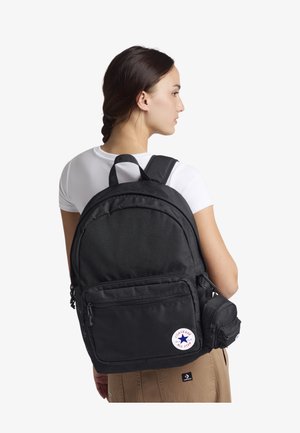 Musthave mustrepp seljakott, mis on valmistatud tekstureeritud kangast, millel on pehmed rihmad, eesmine lukuga tasku, külgkomplekt ja ümmargune logo silt.