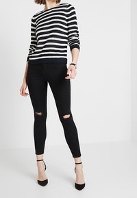 Proužkovaný pletený svetr v námořnické modré a krémové barvě, kombinovaný s černými roztrhanými skinny džíny. Modelka má na sobě černé lodičky na podpatku s pásky kolem kotníků.