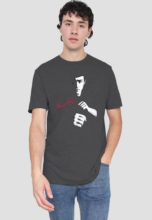 Graues Baumwoll-T-Shirt mit einem bedruckten Grafiken eines Martial-Arts-Kämpfers, dessen Gesicht und Hand in einer Schlagpose zu sehen sind, mit rotem Schriftzug darunter.