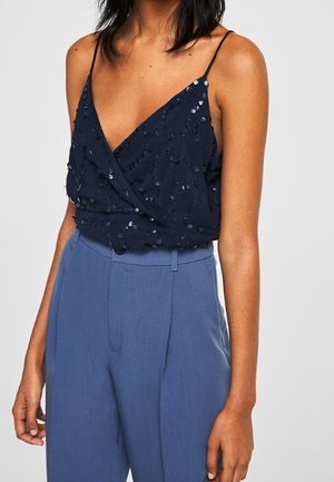 Femme portant un top cache-cœur sans manches bleu marine orné de sequins, associé à un pantalon taille haute bleu clair, ajusté.