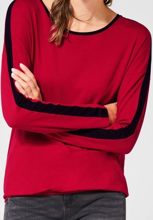 Kobieta ubrana w czerwoną bluzkę z długim rękawem z czarnymi paskami na rękawach, z założonymi skrzyżowanymi ramionami, widać ciemnoszare dżinsy w talii.