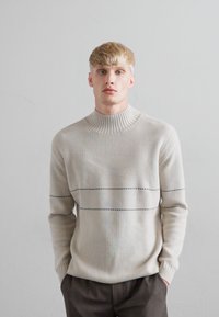 Ung man med blont hår iklädd en beige ribbad tröja med tunna horisontella svarta ränder, stående mot en slät grå vägg.