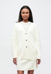 Γυναίκα με σγουρά μαλλιά φοράει ένα λευκό πουλόβερ με κουμπιά πάνω από μια λευκή ριμπ φούστα, στέκεται μπροστά σε έναν απλό ανοιχτό φόντο.